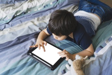 Çocuk ödevi için tablet kullanıyor, çocuk yatakta dijital dokunmatik kâğıtta oyun oynuyor, küçük çocuk tabletle yatıyor. Evde eğitim, sosyal mesafe, e-öğrenim çevrimiçi eğitim.