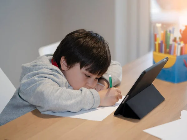 New normal life style, Portrait of school kid sitting alone doing homework, Child boy holding black pen drawing and writing on white paper on table, Elementary school and homeschooling concept