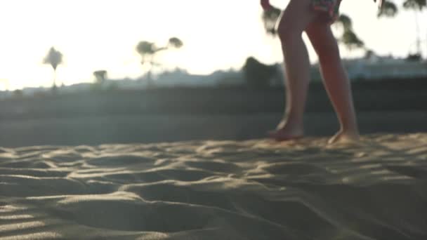 Ένα βίντεο αργής κίνησης μιας νεαρής γυναίκας με καλοκαιρινό φόρεμα να περπατά στην άμμο κατά τη διάρκεια ενός χαμηλού απογευματινού ήλιου — Αρχείο Βίντεο