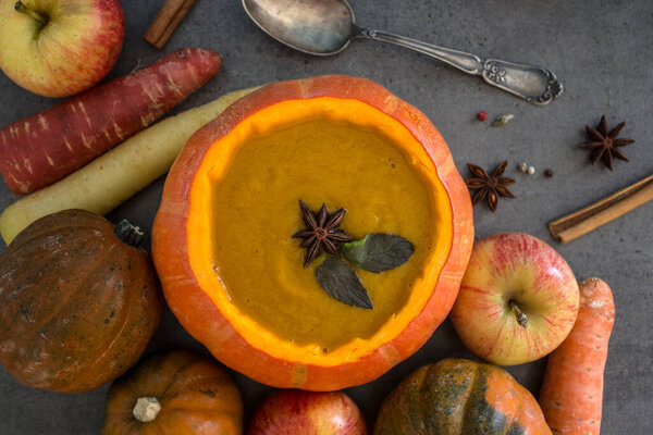 Squash soup in pumpkin bowl on grey textured background with copy space. Top view photo of autumn meal with anise stars, rosemary, cinnamon sticks and silver spoon.
