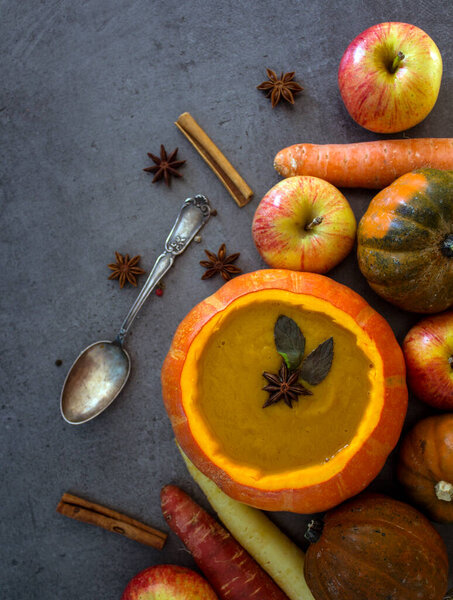 Squash soup in pumpkin bowl on grey textured background with copy space. Top view photo of autumn meal with anise stars, rosemary, cinnamon sticks and silver spoon.