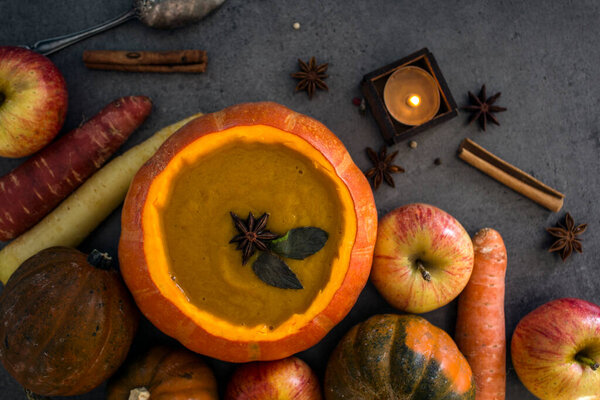 Squash soup in pumpkin bowl on grey textured background with copy space. Top view photo of autumn meal with anise stars, rosemary, cinnamon sticks and silver spoon.