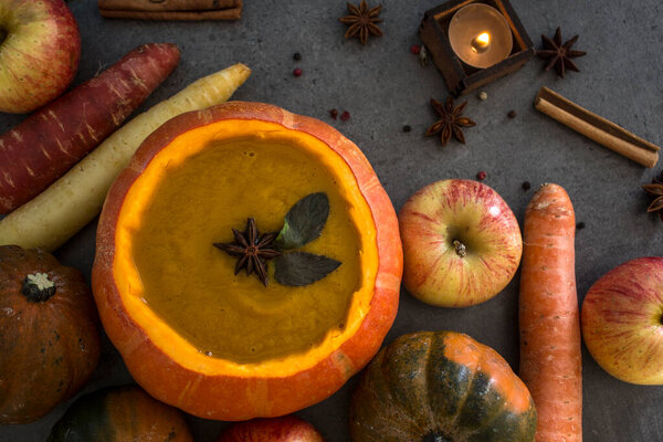 Squash soup in pumpkin bowl on grey textured background with copy space. Top view photo of autumn meal with anise stars, rosemary, cinnamon sticks and silver spoon.