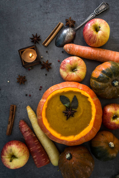 Squash soup in pumpkin bowl on grey textured background with copy space. Top view photo of autumn meal with anise stars, rosemary, cinnamon sticks and silver spoon.