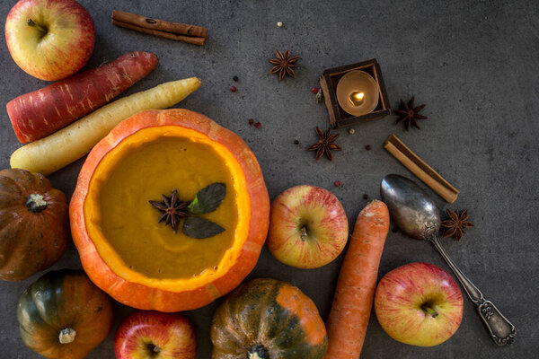 Squash soup in pumpkin bowl on grey textured background with copy space. Top view photo of autumn meal with anise stars, rosemary, cinnamon sticks and silver spoon.