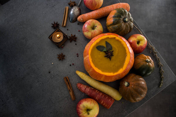 Squash soup in pumpkin bowl on grey textured background with copy space. Top view photo of autumn meal with anise stars, rosemary, cinnamon sticks and silver spoon.