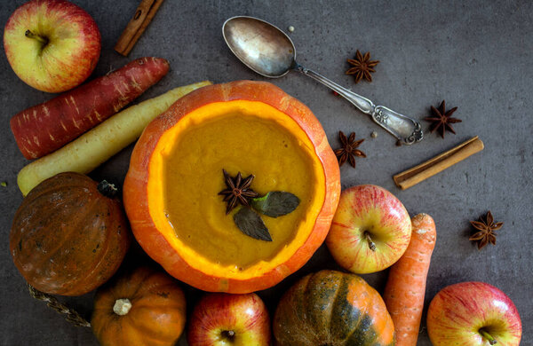 Squash soup in pumpkin bowl. Top view photo of pumpkin, apples, carrots anise stars and cinnamon sticks. Grey background.Autumn food close up photo. 