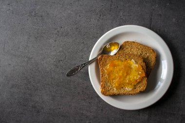 Beyaz tabakta tereyağlı ve reçelli tost resmi. Koyu gri desenli arka plan. 
