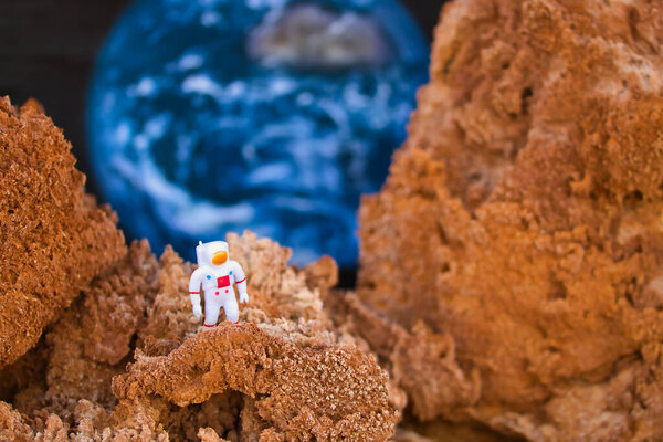 Toy astronaut exploring the mountains of planet Mars with the Chinese flag on the space suit and the earth behind.
