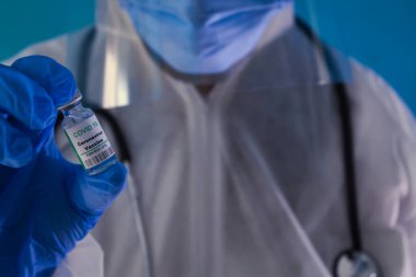 Mavi eldivenli, maskeli, gözlüklü, steteskoplu yeşil bir aşı taşıyan doktor..