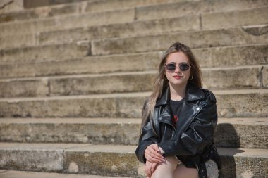 Genç bir kadın, güzel, sarışın, deri ceketli, yılan etekli, güneş gözlüklü, kameraya bakan, merdivenlerde oturan. İsyankar, boyun eğmez, isyancı konsept.