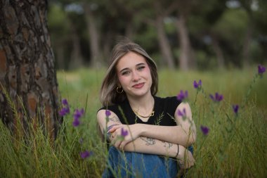 Genç bir kadın, güzel, sarışın, yeşil gözlü, siyah tişört ve kot pantolon giyen, gülümseyen ve mutlu, çimenlerde oturan ve mor çiçeklerle çevrili. Mutluluk kavramı, çiçekler, bahar.