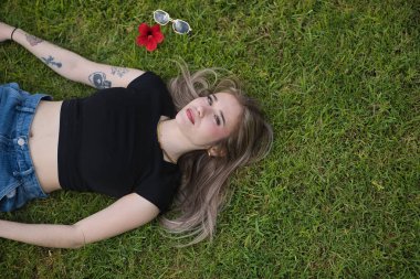 Genç bir kadın, güzel, sarışın, yeşil gözlü, rahat ve sakin, yukarıdan görülüyor, kameraya bakıyor ve yeşil çimlerin üzerinde yatıyor. Rahatlama, huzur, bahar, çimen kavramı.