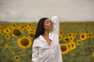 Genç, güzel, Latin esmer kadın beyaz gömlek giyiyor, gözleri kapalı, doğaya bağlı, ayçiçeği tarlasının ortasında. Kavram: doğa, tarım, kırsal.