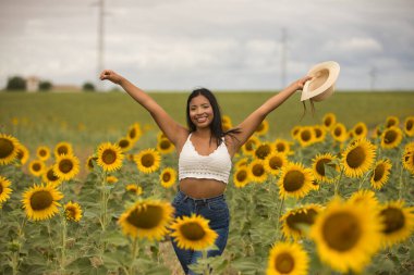 Genç, çekici, latin esmer kadın beyaz örgü örer, kot pantolon giyer ve elinde bir şapka, kolları ayçiçeği tarlasında mutlu bir şekilde gülümserdi. Kavram: doğa, tarım.