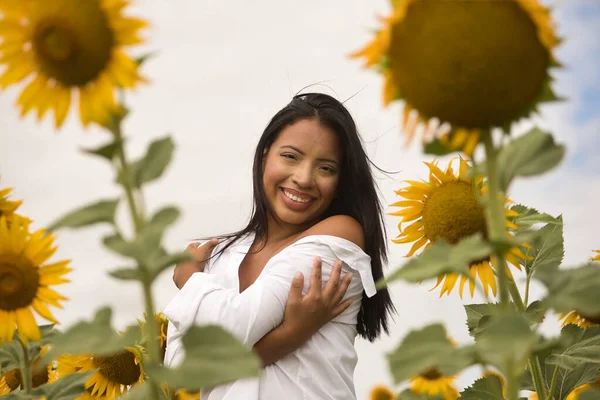 Beyaz gömlek giyen, kendine sarılan, gülümseyen ve mutlu bir Latin esmer kadının portresi, bir ayçiçeği tarlasının bitkilerinin arasından görülüyor. Kavram: doğa, tarım.