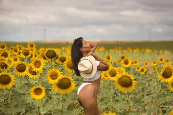 Genç, güzel, Latin esmer kadın beyaz bir bluz ve bikini giyiyor, şapka, şehvetli ve kışkırtıcı, ayçiçeği tarlasının ortasında. Kavram: doğa, tarım, kırsal.