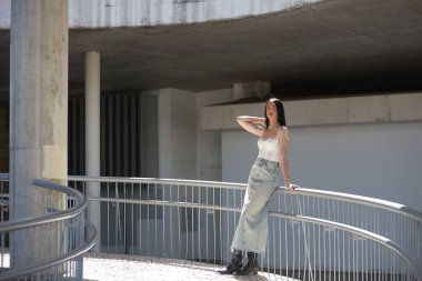 Genç bir kadın, esmer, uzun boylu, zayıf ve çok dövmeli, beyaz bluzlu, kot etekli ve botlu, kameraya bakıyor, saçlarına güneş ışınları tutuyor, korkuluklara yaslanıyor..