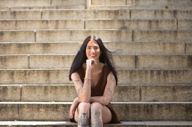 Genç bir kadın, koyu saçlı, uzun boylu, zayıf ve çok dövmeli, kahverengi bir elbise giyiyor, eli çenesinde, kameraya bakıyor, gülümsüyor, mutlu, masum ve yalnız, merdivenlerde oturuyor..