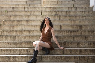 Genç bir kadın, esmer, uzun boylu, zayıf ve çok dövmeli, kahverengi elbiseli ve çizmeli, tatlı, masum ve yalnız, merdivenlerde oturuyor. Gençlik kavramı, güzellik, asilik, milenyum.