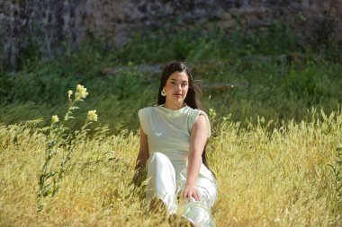 Beyaz elbiseli, genç, güzel, esmer kadın, kameraya bakıyor, rahat ve sakin, rüzgarlı bir günde uzun otların arasında oturuyor. Güzellik, moda, moda, gençlik kavramı.