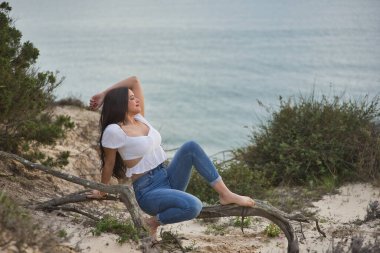 Genç, güzel bir kadın, siyah saçlı, beyaz bir gömlek giyiyor, kot pantolon giyiyor, rahat ve sakin, denize bakıyor, ağacın kökünde oturuyor. Güzelliği, modayı, doğayı, kumsalı, denizi kavra.