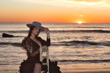 Kadın, genç, güzel, esmer, flamenko dansçısı, kordova şapkalı ve çiçekli siyah flamenko elbiseli, plajda arka planda deniz olan bir sandalyede oturuyor..