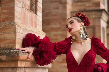 Genç, güzel, kırmızı flamenko elbiseli sarışın kadın, saçında çiçekler ve altın mücevherler, Seville 'de İspanya meydanında tuğla bir duvara yaslanmış poz veriyor. Dans konsepti, sanat, tipik İspanyol..