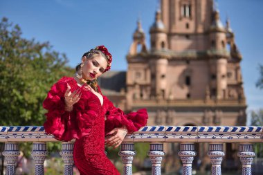 Genç, güzel, sarışın, kırmızı flamenko elbiseli, saçında çiçekler ve altın mücevherlerle, Seville 'de İspanya meydanında fayanslı bir korkuluğa yaslanmış flamenko pozları veriyor. Dans konsepti, sanat.