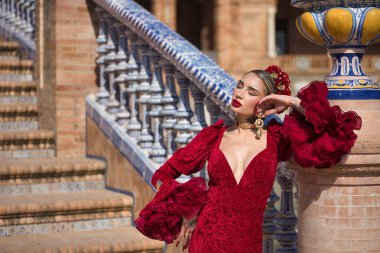 Genç, güzel, sarışın, kırmızı flamenko elbiseli, saçında çiçekler ve altın mücevherlerle, İspanya 'nın Seville meydanında bir tuğla sütuna yaslanmış flamenko pozlar veriyor. Dans konsepti, sanat.
