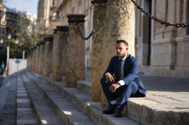 Genç, esmer, İspanyol, modern yönetici, takım elbiseli ve kravatlı, ciddi, ekonomik krizden endişeli ve sıkıntılı, bir şehir caddesinde bazı basamaklarda oturuyor. İş konsepti, şirket, lider.