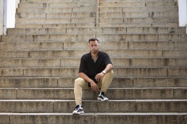 Genç, esmer, İspanyol, siyah tığ işi gömlek ve kot pantolon giyen, yalnız bir merdivenin ortasında sigara içen modern bir adam. Moda, moda, gündelik konsept.