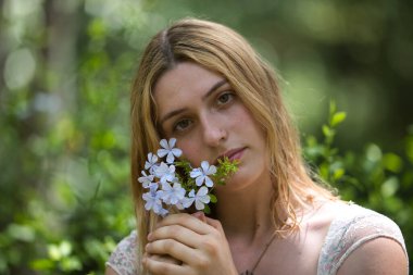 Genç bir kadının portresi, güzel, sarışın, çiçekli bir elbise giymiş, tatlı ve narin bakışlı, yüzünün yanında bir buket menekşe tutan, açık hava parkında. Güzellik kavramı, bin yıllık.