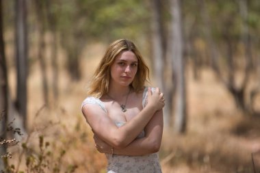 Genç, güzel, sarışın bir kadın, çiçekli bir elbise giyiyor, kendini kucaklıyor, kendini seviyor ve bir koruma işaretiyle, açık hava parkında. Güzellik kavramı, gençlik, milenyum.