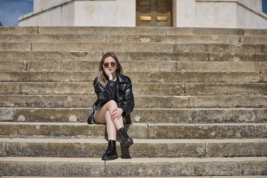 Genç, güzel, sarışın, deri ceketli, yılan etekli, çizmeli ve güneş gözlüklü, rahat ve sakin bir şekilde merdivende oturan kameraya bakıyor. İsyankar, boyun eğmez bir kavram..