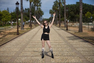 Genç, güzel, sarışın bir kadın, siyah bluzlu ve etekli, botları ve güneş gözlüklü, kolları havada, mutlu ve hayattan memnun, bir caddenin ortasında. Minnettarlığı, mutluluğu, minnettarlığı kabul et..