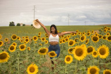 Genç, çekici, latin esmer kadın beyaz örgü örer, kot pantolon giyer ve elinde bir şapka, kolları ayçiçeği tarlasında mutlu bir şekilde gülümserdi. Kavram: doğa, tarım.