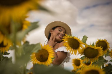 Beyaz bir silindir ve şapka giyen güzel Latin esmer bir kadının portresi, ayçiçeği tarlasında bitkilerin arasında kendini kucaklıyor ve seviyor. Kavram: doğa, tarım, kırsal.