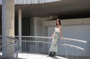 Genç bir kadın, esmer, uzun boylu, zayıf ve çok dövmeli, beyaz bluzlu, kot etekli ve botlu, kameraya bakıyor, saçlarına güneş ışınları tutuyor, korkuluklara yaslanıyor..