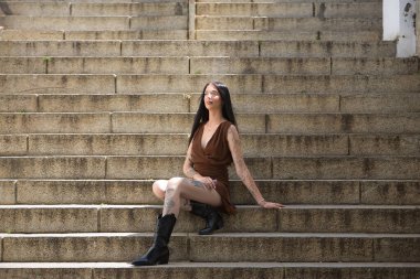 Genç bir kadın, esmer, uzun boylu, zayıf ve çok dövmeli, kahverengi elbiseli ve çizmeli, tatlı, masum ve yalnız, buharlaşmış, merdivenlerde oturuyor. Gençlik kavramı, güzellik, asilik, milenyum.