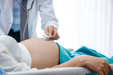 Doktor, klinikteki yatakta yatan bir kadının hamileliğini kontrol etmek için çan şeklinde stetoskop kullanmış..