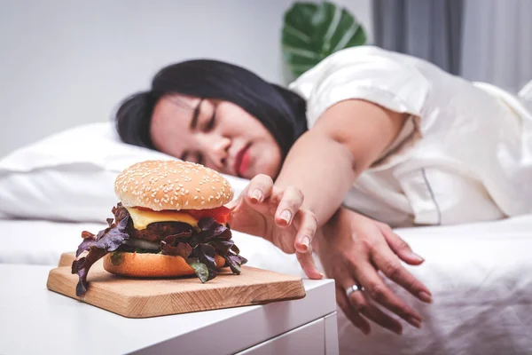 Wanita Gemuk Berbaring Tempat Tidur Mencapai Hamburger Kosong Atas Meja — Stok Foto