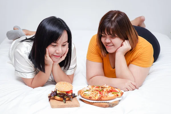 Two fat girls eat pizza and hamburgers in bed in the bedroom. Happy. The concept of eating and health care.