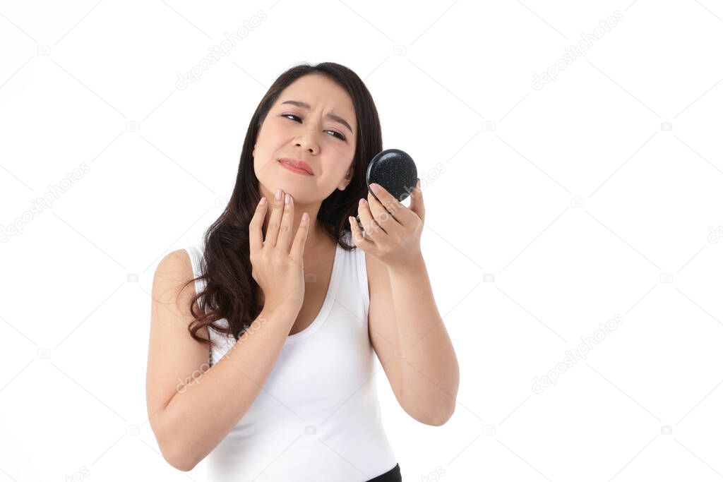 A beautiful Asian woman feels stressed. She is holding a mirror, looking at acne on her cheeks. Beauty concept, white background