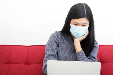 Maske takan Asyalı bir kadın oturma odasında oturup çalışıyor. Sosyal mesafe kavramı. Yeni normal
