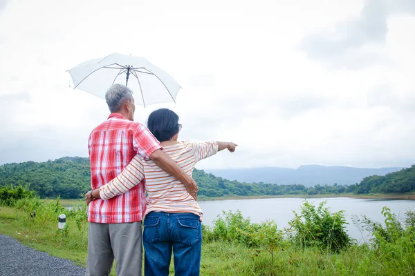 年事已高的恋人站在一起 丈夫为妻子拿着雨伞 是一个可爱的夫妇站在那里观看国家雨季的山景 老年人社区概念 — 图库照片