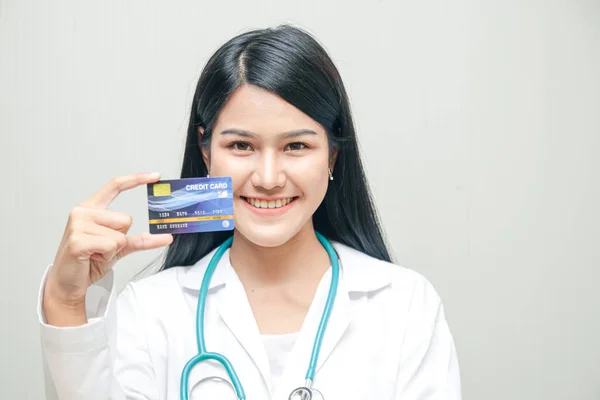 Beautiful Asian doctor holding a credit card standing smiling brightly White backdrop. Health service concept And finance in one card