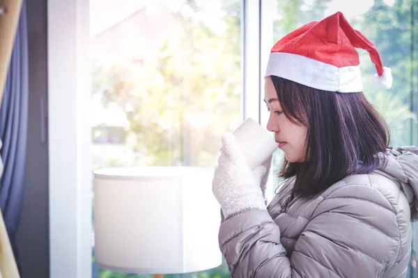 Woman wearing a red Christmas hat, wearing gloves and holding a white glass, standing by the window. Festival concept