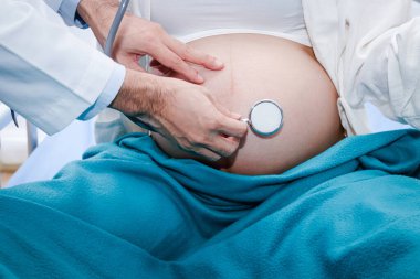 Doktor, klinikteki yatakta yatan kadının hamileliğini kontrol etmek için steteskop kullanıyor..
