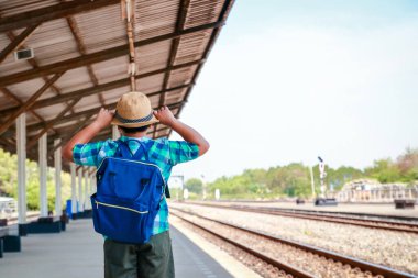 Asyalı küçük bir çocuk mavi bir sırt çantası taşıyor. Trenin okula gitmesini bekliyor.